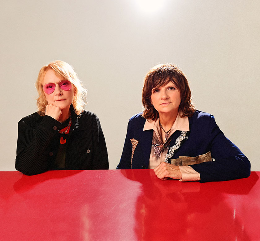 Image of the two members of Indigo Girls at a red table with a white background.